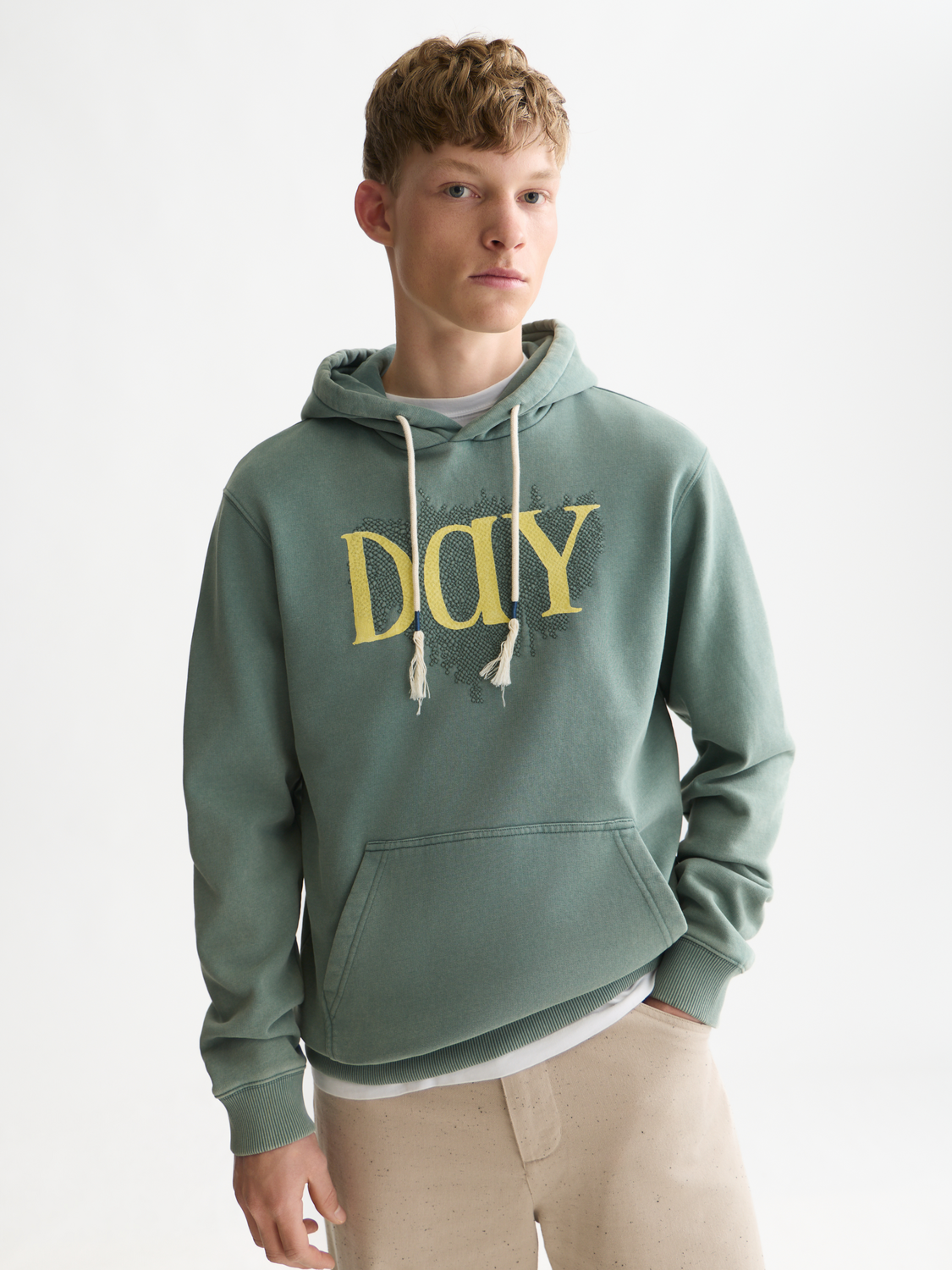 A young man wearing a sage green pullover hoodie with a distressed yellow 'DAY' graphic on the chest, styled with beige pants against a clean white studio background.