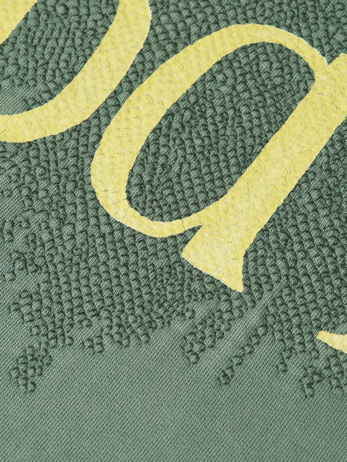 Close-up of a green knit fabric with a large pale yellow letter or graphic element partially visible.