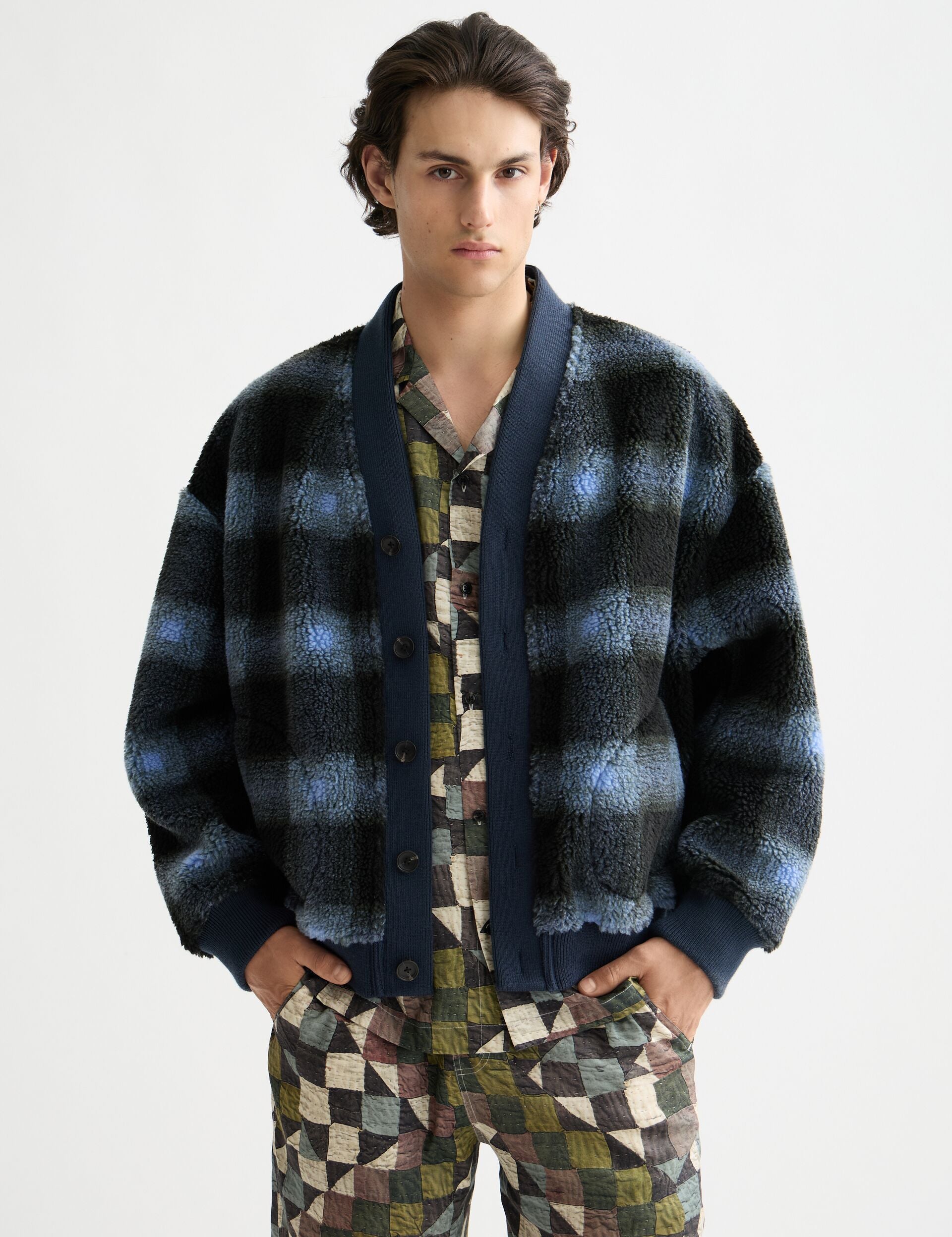 Person wearing a plaid cardigan over a patterned shirt and pants on a light gray background